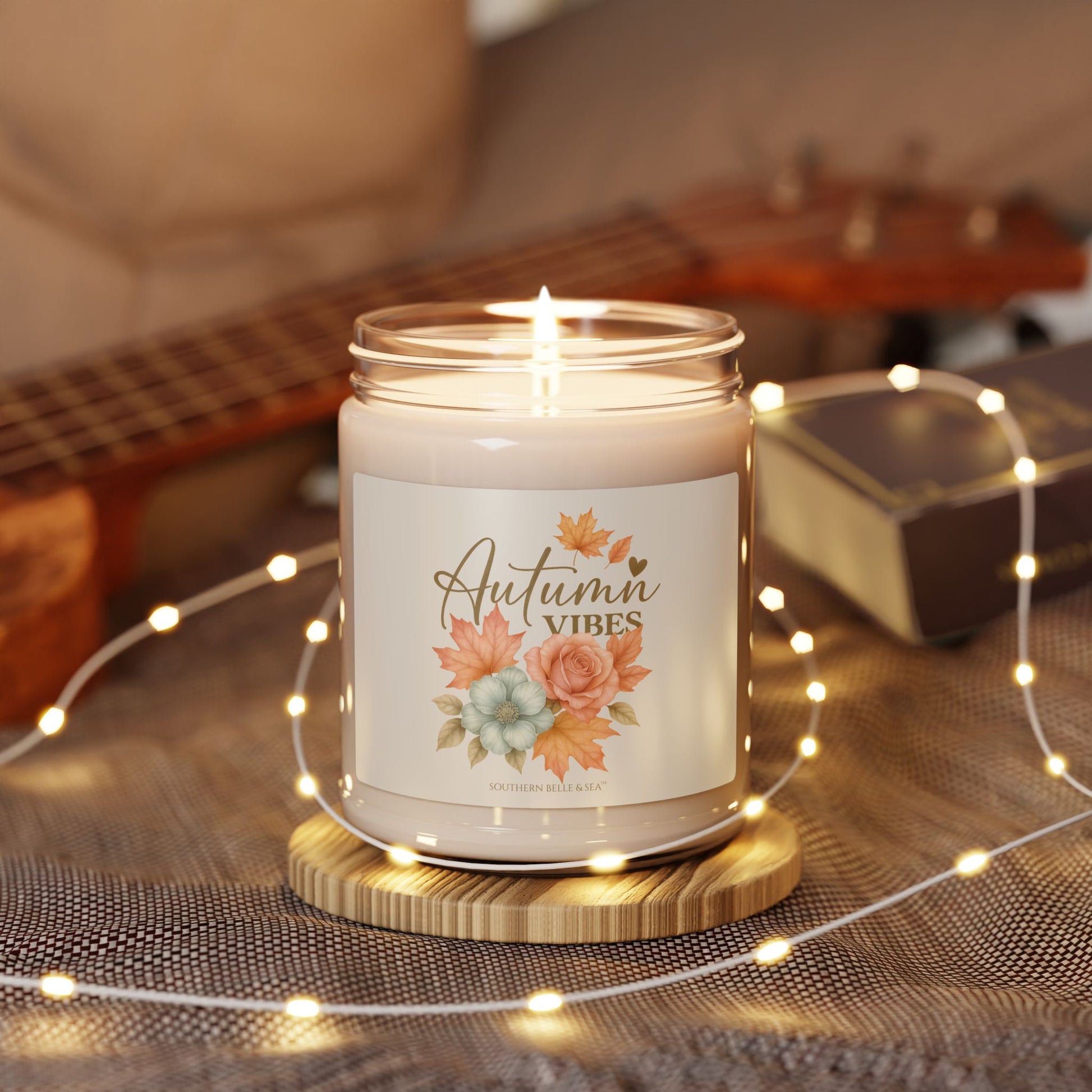 Candle with 'Autumn Vibes' label on a wooden coaster surrounded by string lights and a guitar in the background.