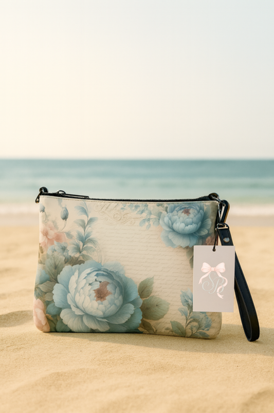 Southern Belle & Sea™ blue floral crossbody bag resting on the sand at the beach with soft pastel flowers and black strap.