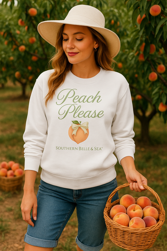 Woman in a peach orchard wearing a white “Peach Please” sweatshirt from Southern Belle & Sea™, holding a wicker basket filled with fresh peaches and wearing a wide-brimmed hat.