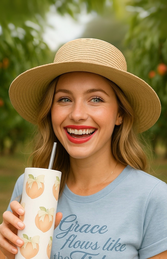Woman in straw hat holding the Peach Please stainless tumbler with peach motif, smiling in an orchard.