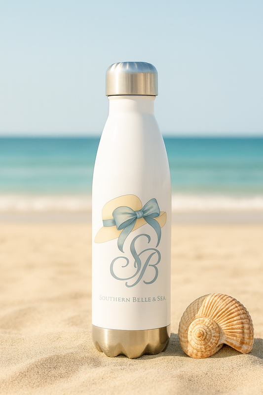 Elegant stainless steel water bottle featuring the Southern Belle & Sea™ logo with a vintage sunhat and powder blue bow, standing upright in soft beach sand next to a spiral seashell, with ocean waves and clear blue sky in the background.

