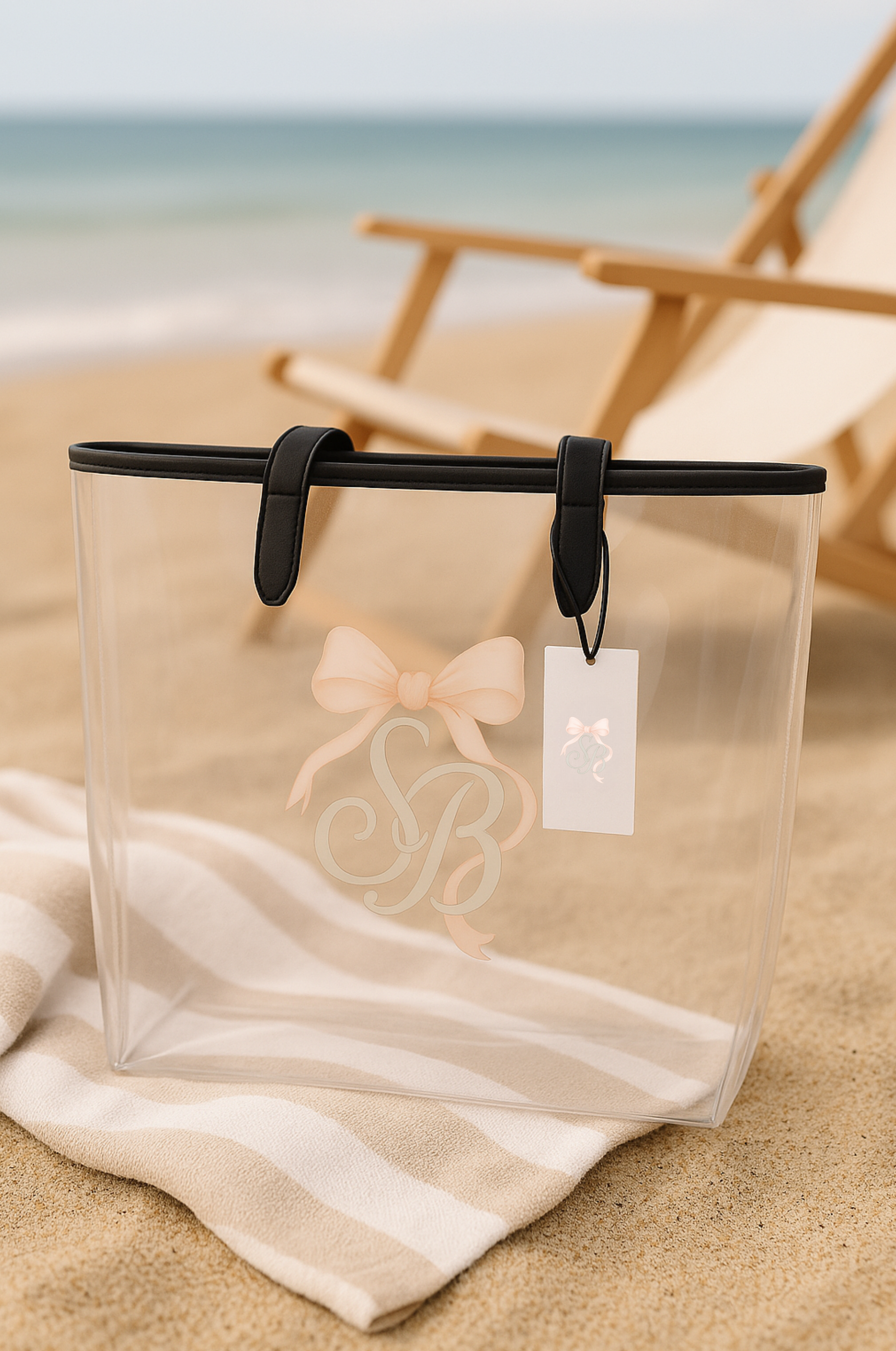 Elegant Southern Belle clear tote bag with blush bow monogram resting on striped beach towel by the ocean — perfect coastal accessory for beach days, summer travel, or minimalist resort fashion.